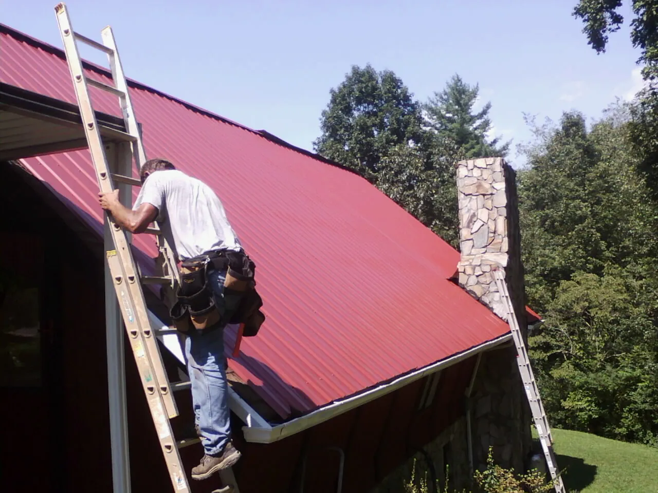 Roofer on a ladder beside a red metal roof and stone chimney — Soffit & Fascia Repair by Roofing Brothers in Five Points